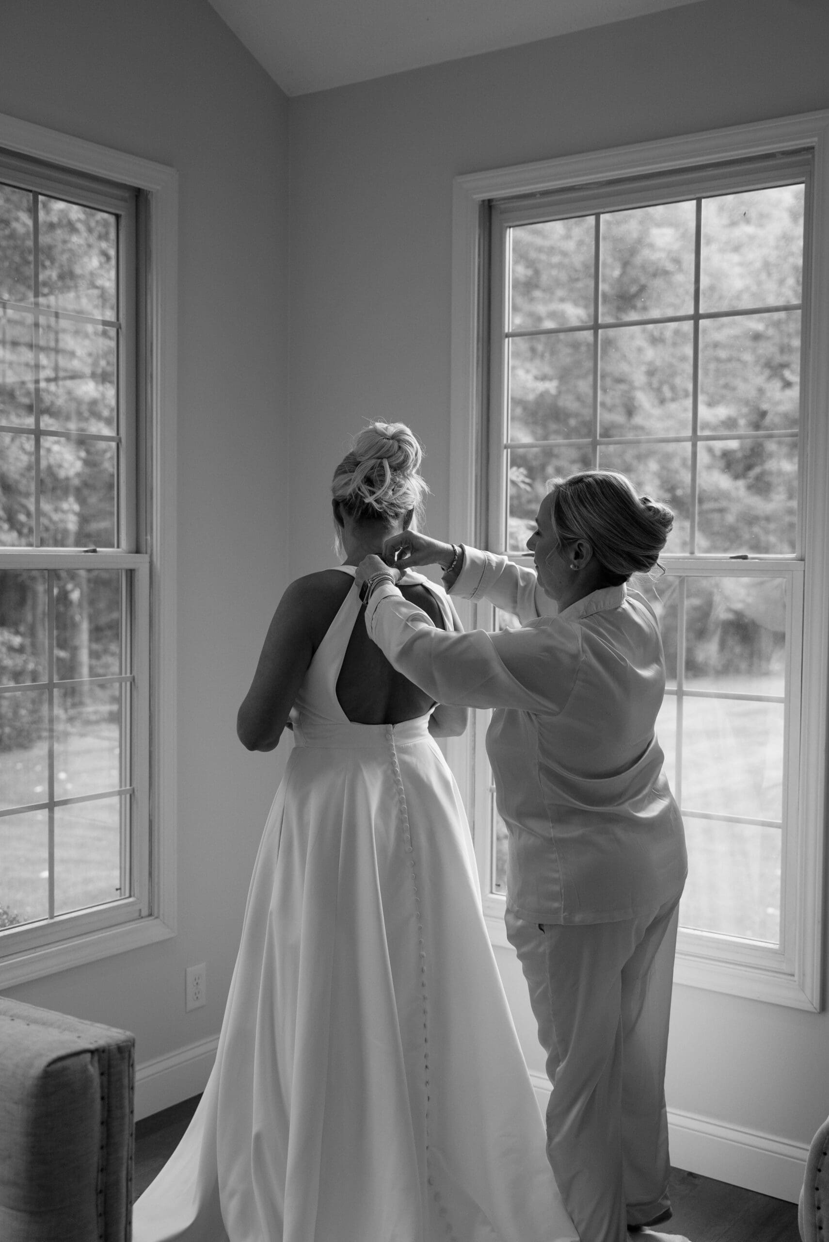 A black and white photo of a bride having her wedding gown fastened by a family member in a bright bridal suite with large natural light windows in Iowa.