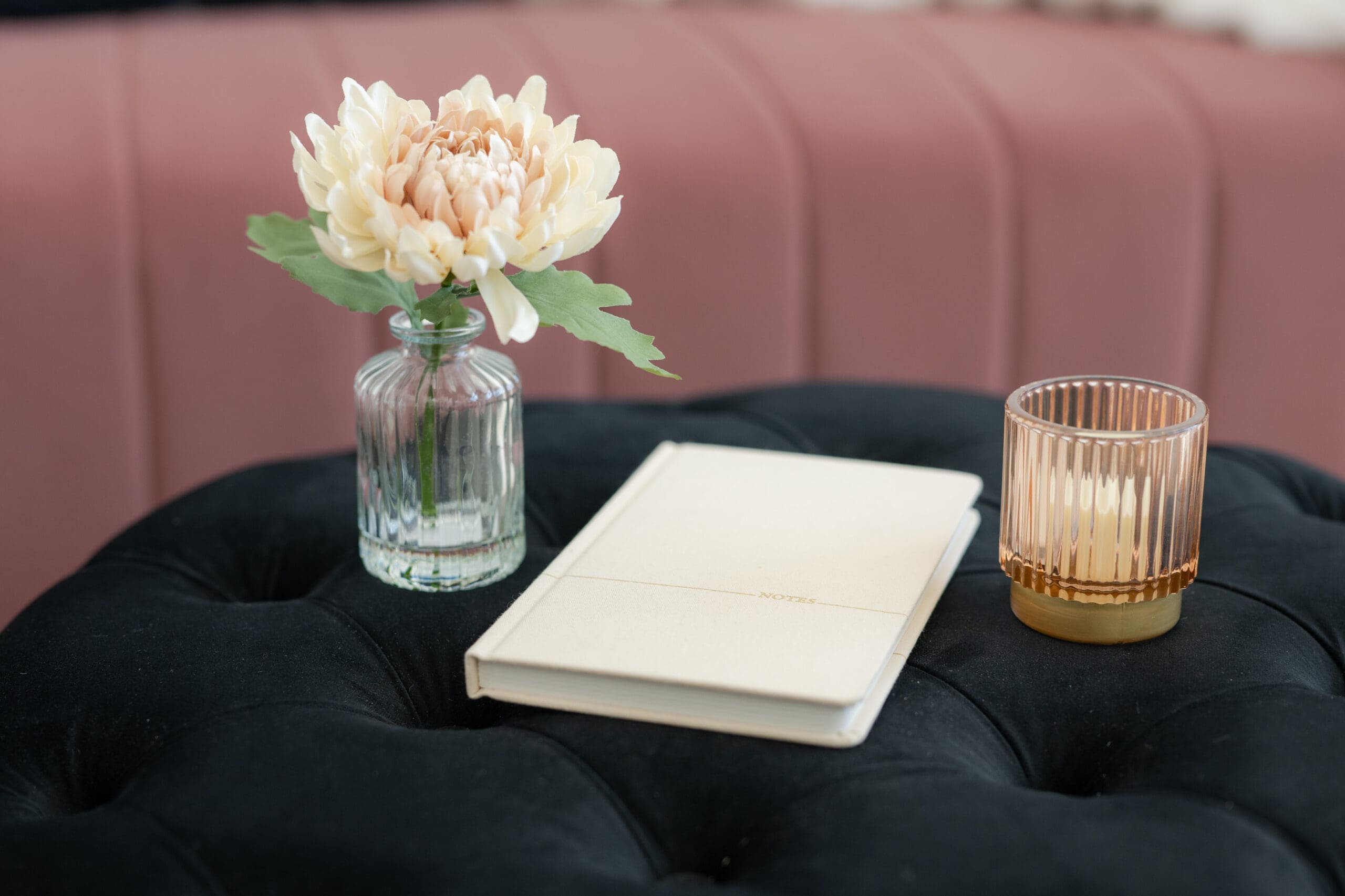 A tufted black velvet ottoman styled with a peach chrysanthemum in a glass vase, a cream notebook, and an amber candle holder in a bridal suite lounge.