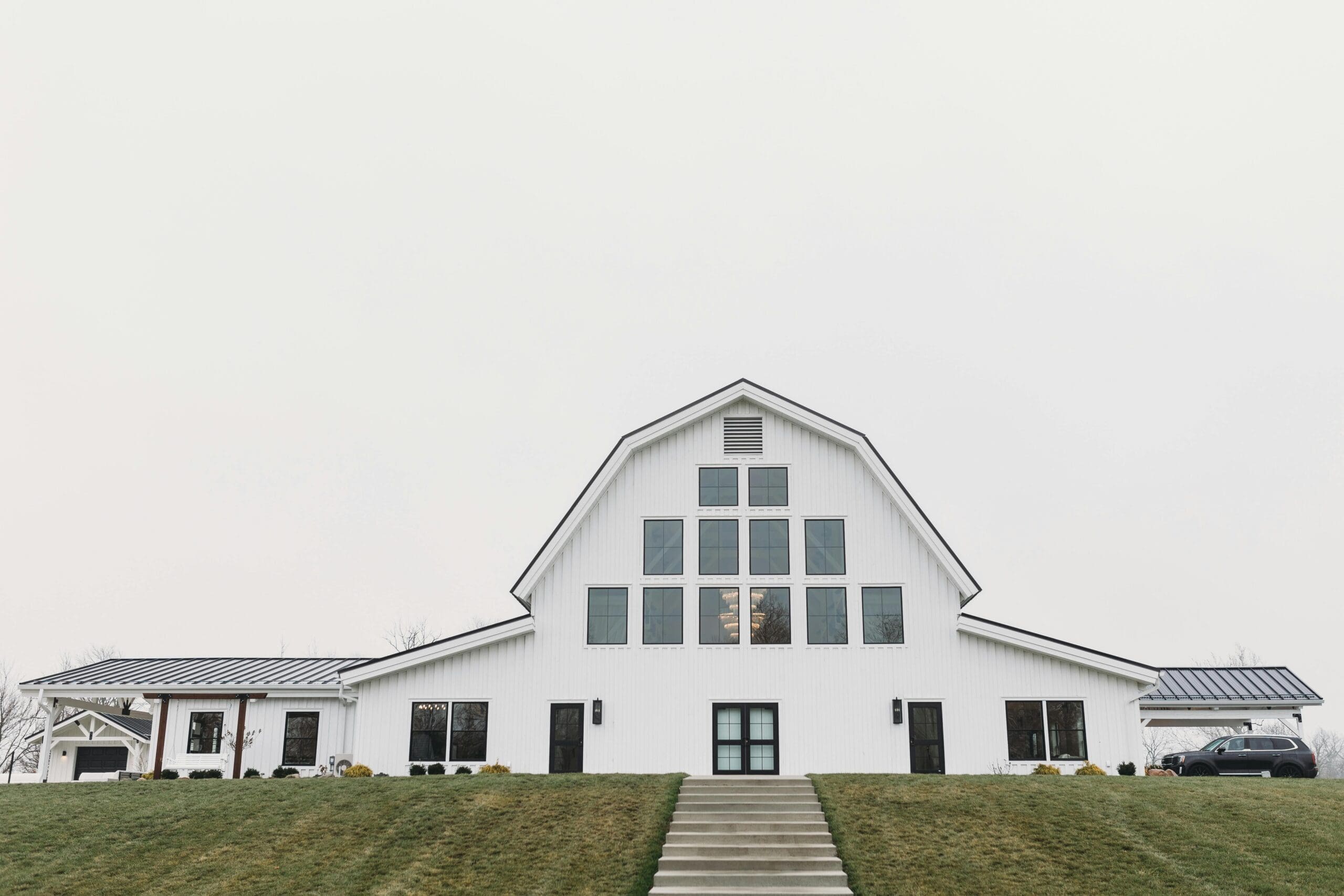 A white modern barn wedding venue exterior with floor-to-ceiling windows, black trim, and a grand central entrance on a green hillside in Iowa.