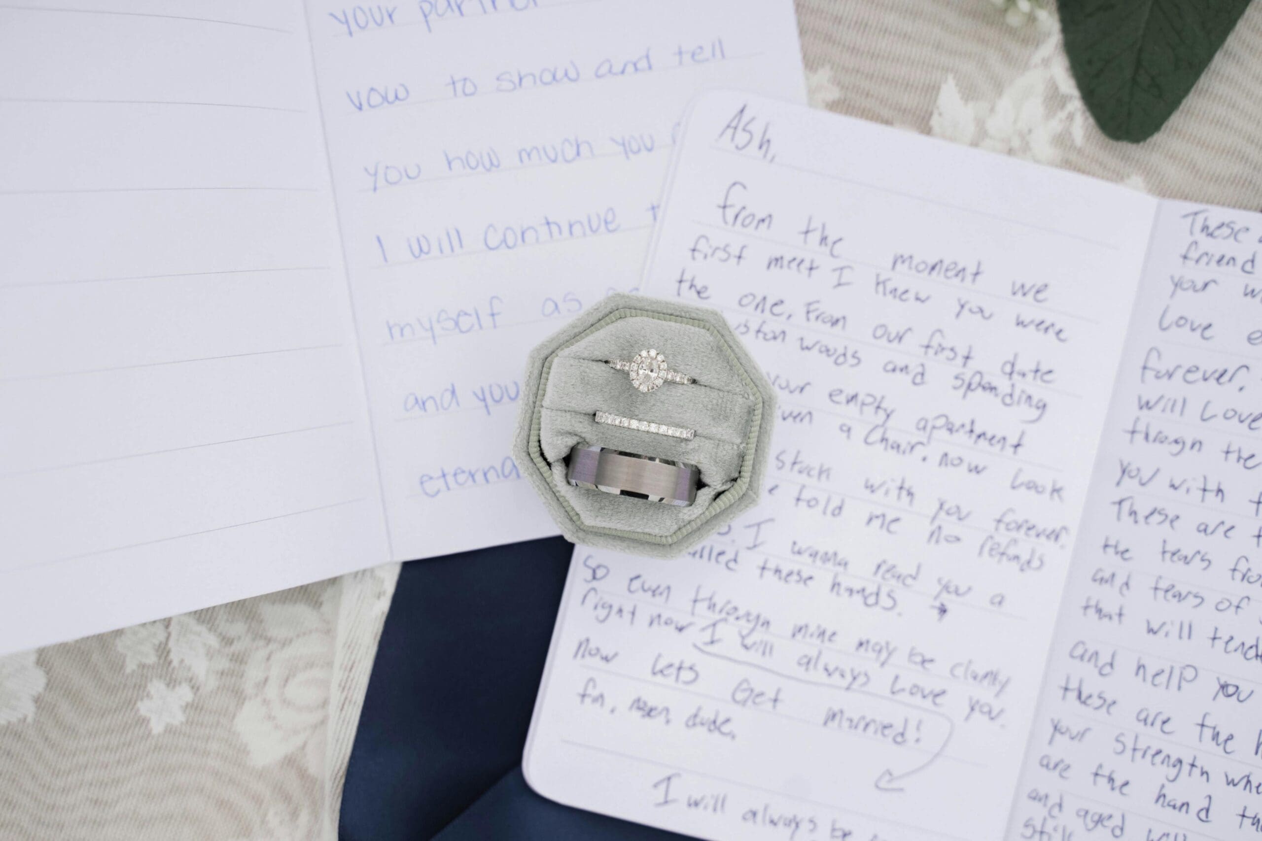 A sage green velvet ring box with an oval diamond engagement ring, wedding band, and groom's band resting on top of handwritten wedding vows.