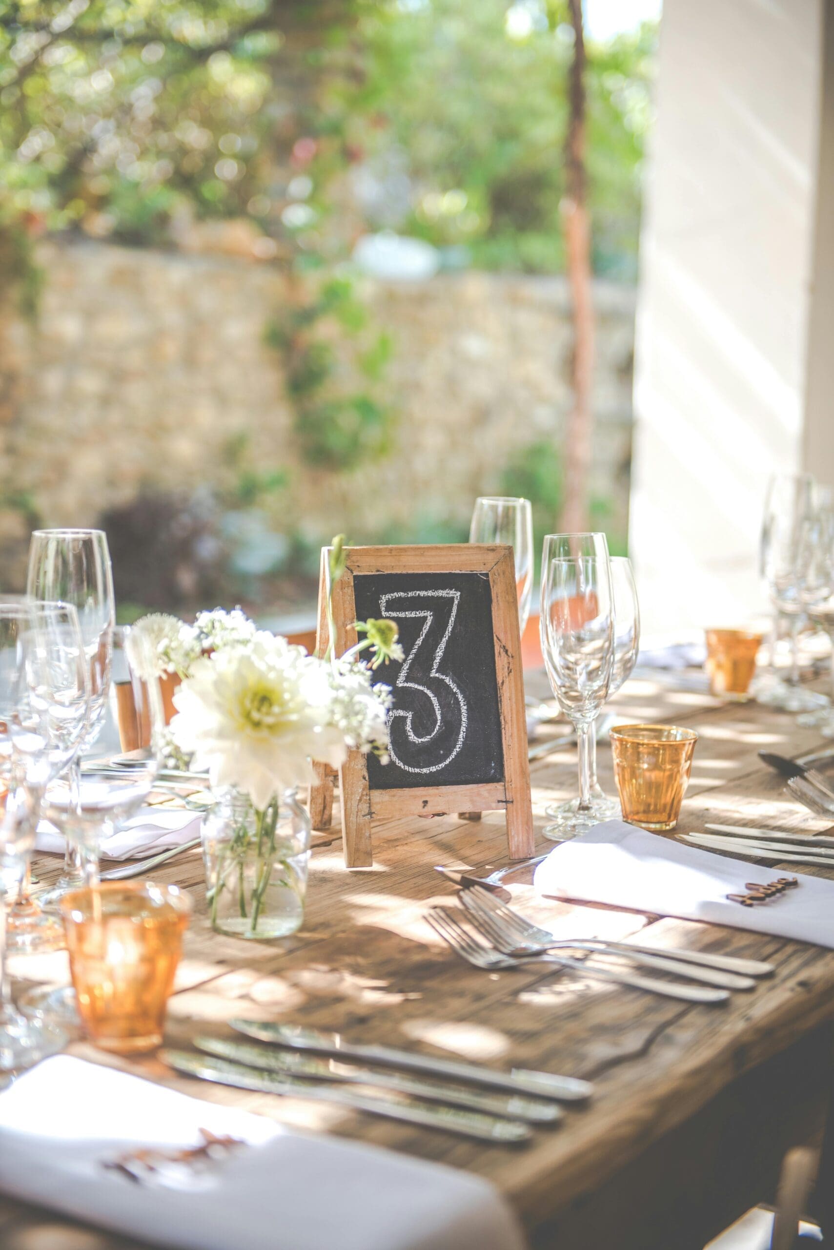 A sunlit rustic wedding reception table set with white florals, champagne flutes, amber glassware, and a chalkboard table number at an outdoor Iowa wedding venue.