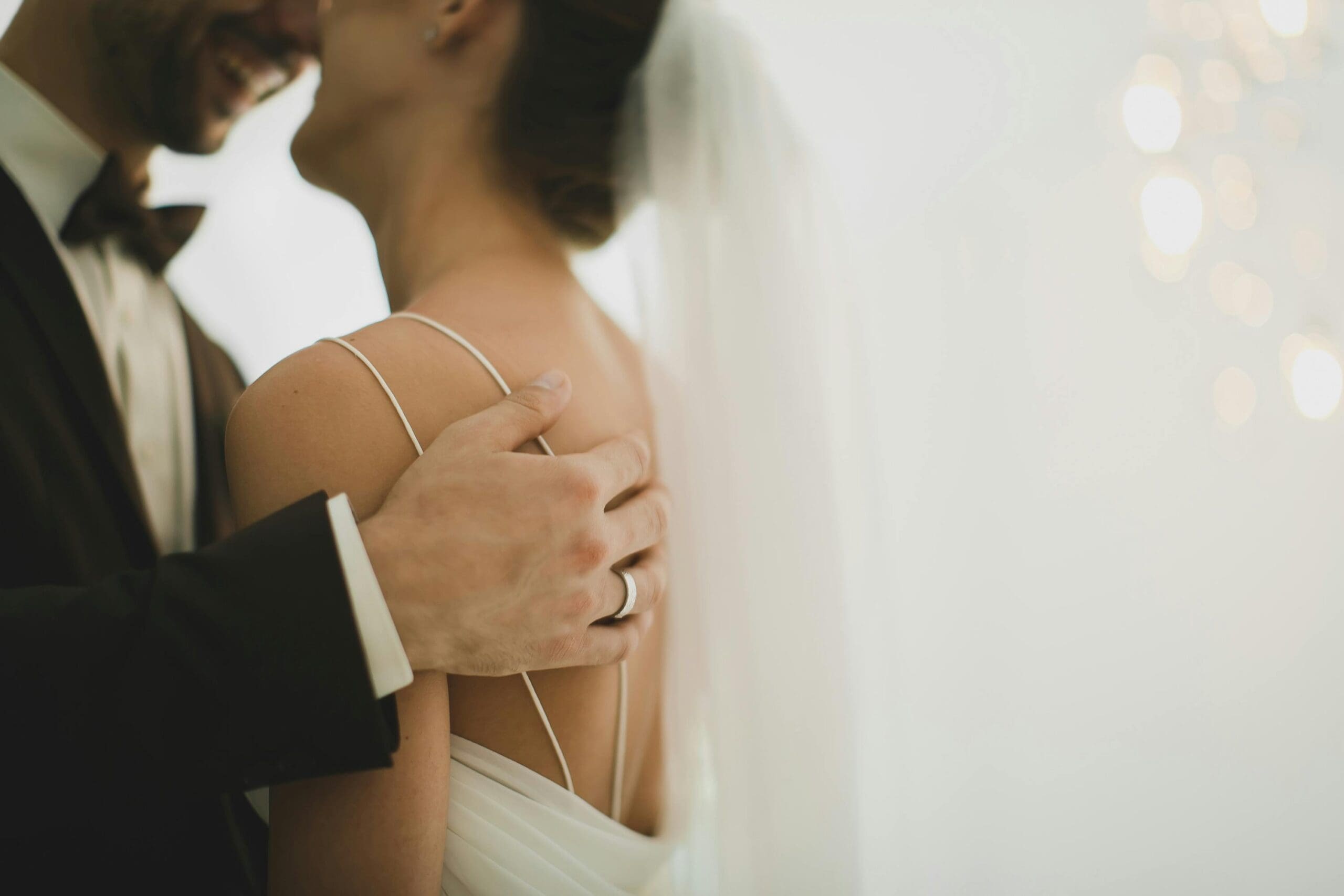 A groom in a black tuxedo holding a bride's shoulder during an intimate wedding portrait, with soft bokeh light and a flowing veil in the background.