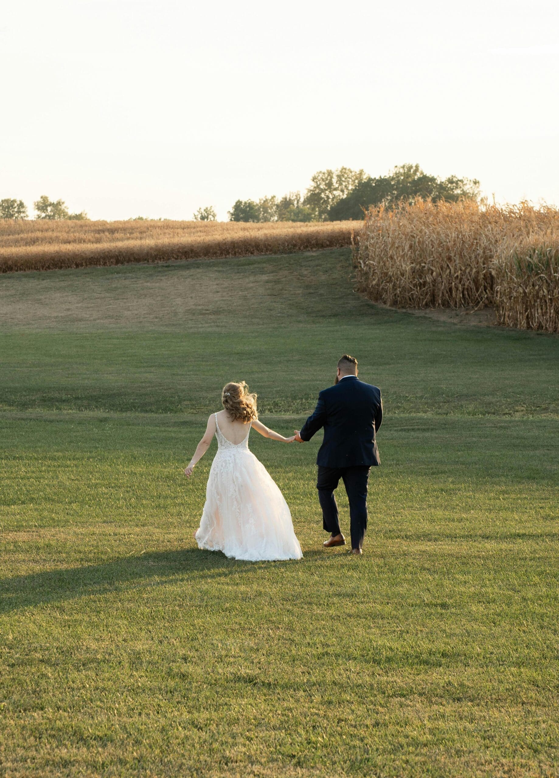A bride in a lace backless gown and groom in a navy suit walking hand in hand through a golden Iowa field at sunset during wedding portraits.