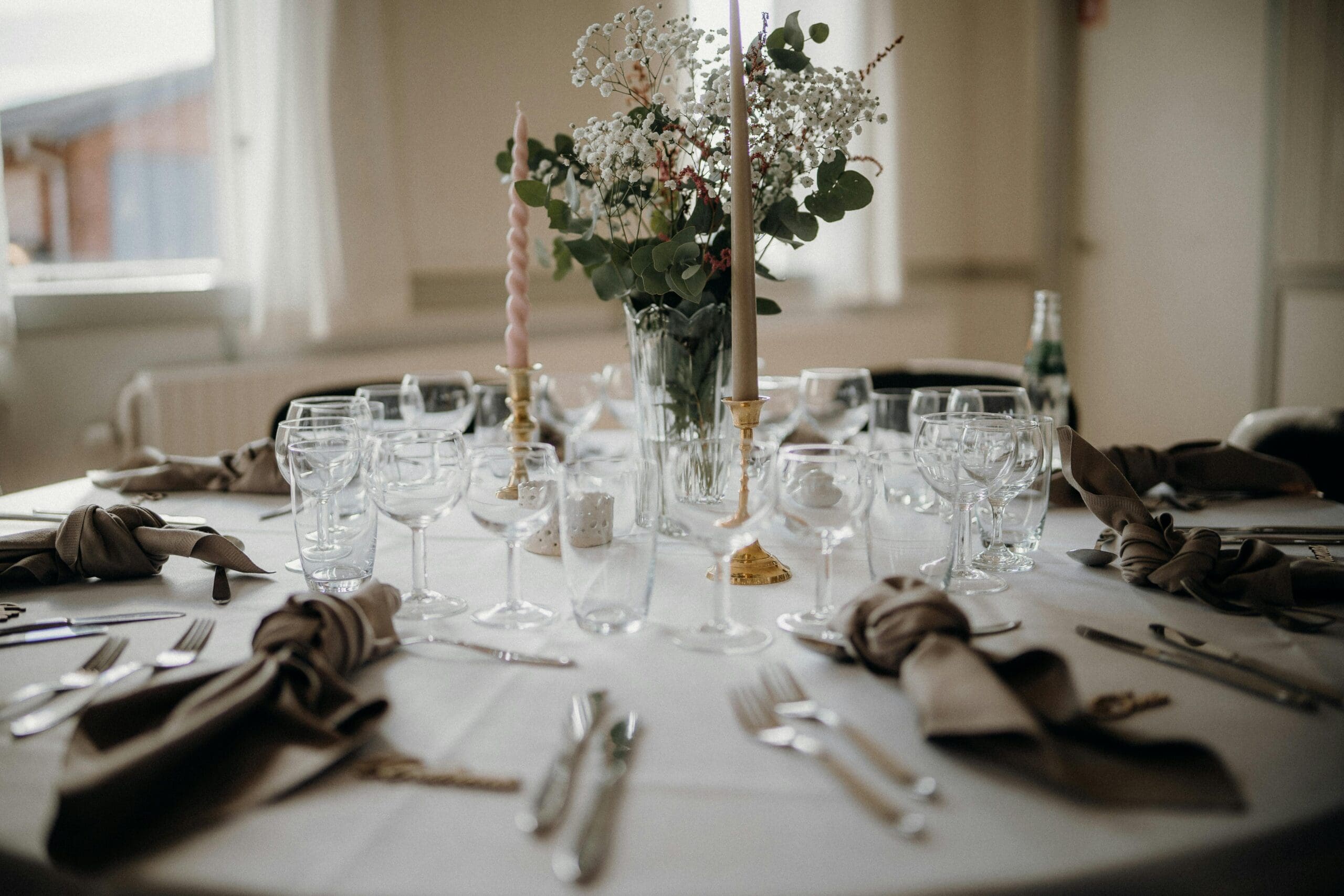 A wedding reception table set with taupe linen napkins, crystal glassware, gold candlesticks, and a wildflower centerpiece in a bright Iowa wedding venue.