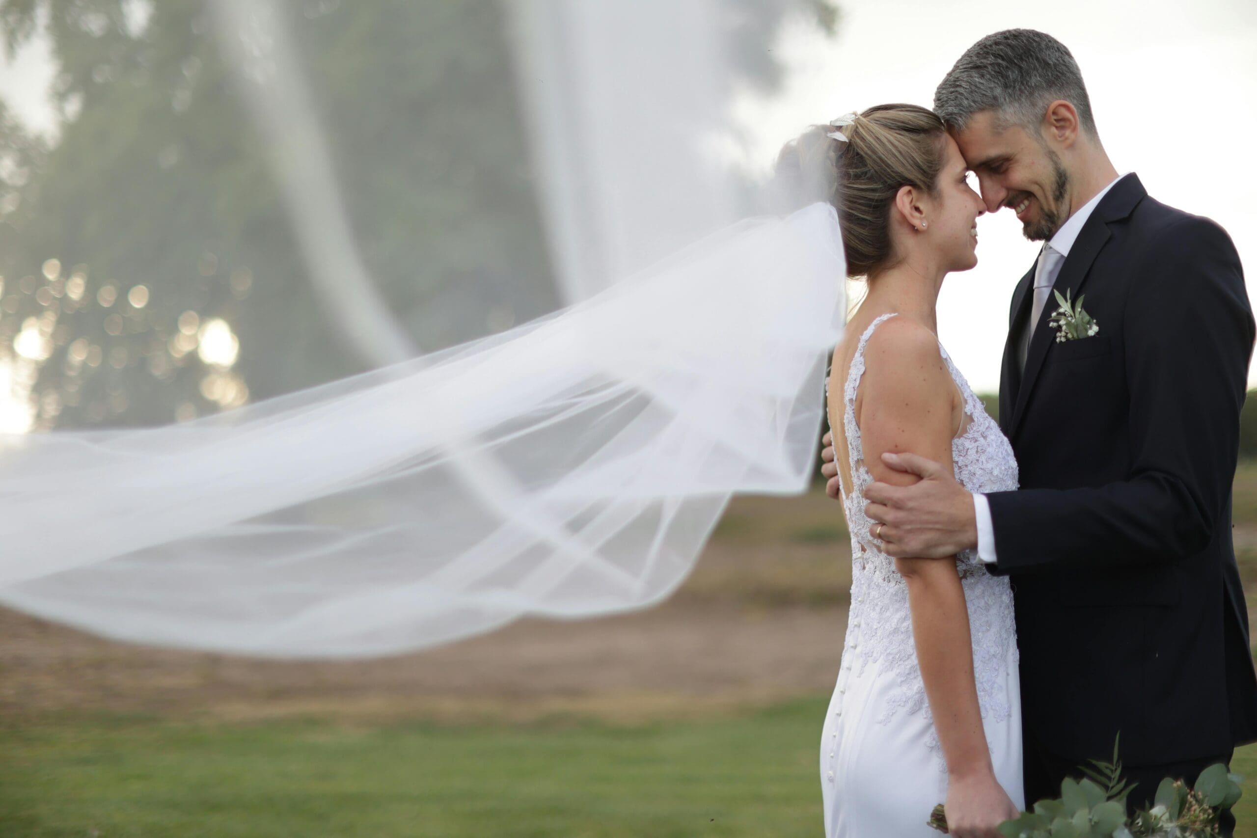 A bride and groom touching foreheads and smiling during outdoor wedding portraits, the bride's veil flowing dramatically in the wind against a soft misty background.