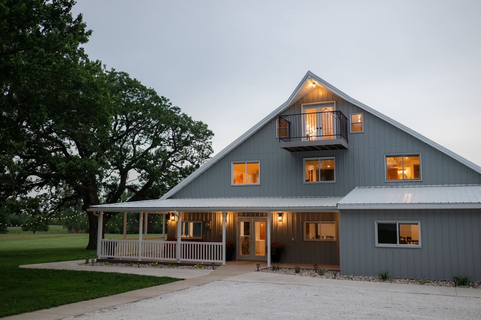 The Era wedding venue exterior at dusk, a gray barn-style building with warm glowing windows, a wraparound porch, string lights, and a private balcony overlooking green grounds in Iowa.