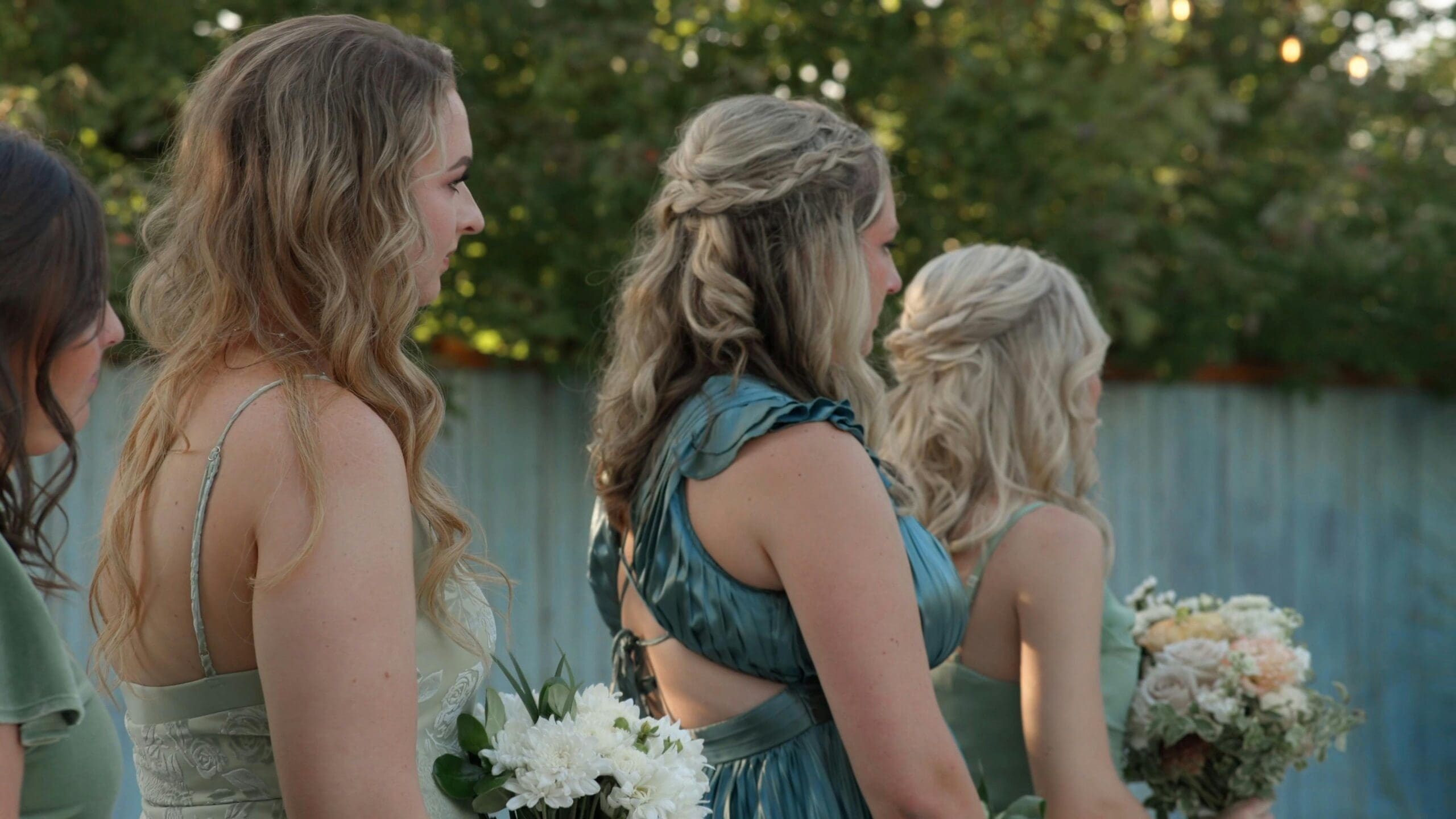 Bridesmaids in sage and teal gowns holding white and blush bouquets, standing together at an outdoor wedding ceremony at golden hour.