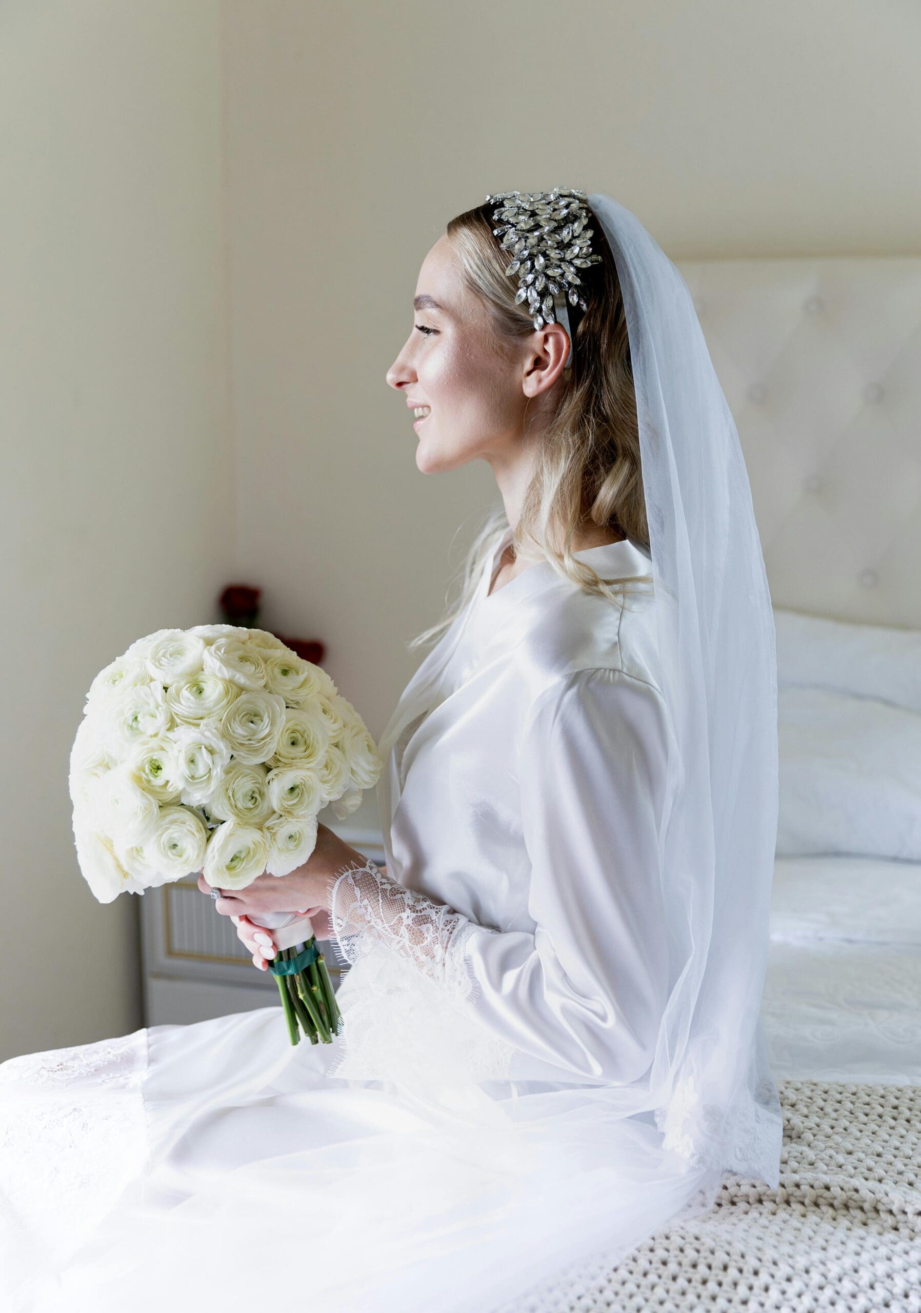 A bride in a white silk robe and veil seated on a bed holding a white ranunculus bouquet, wearing a crystal headpiece, in a bright bridal suite.
