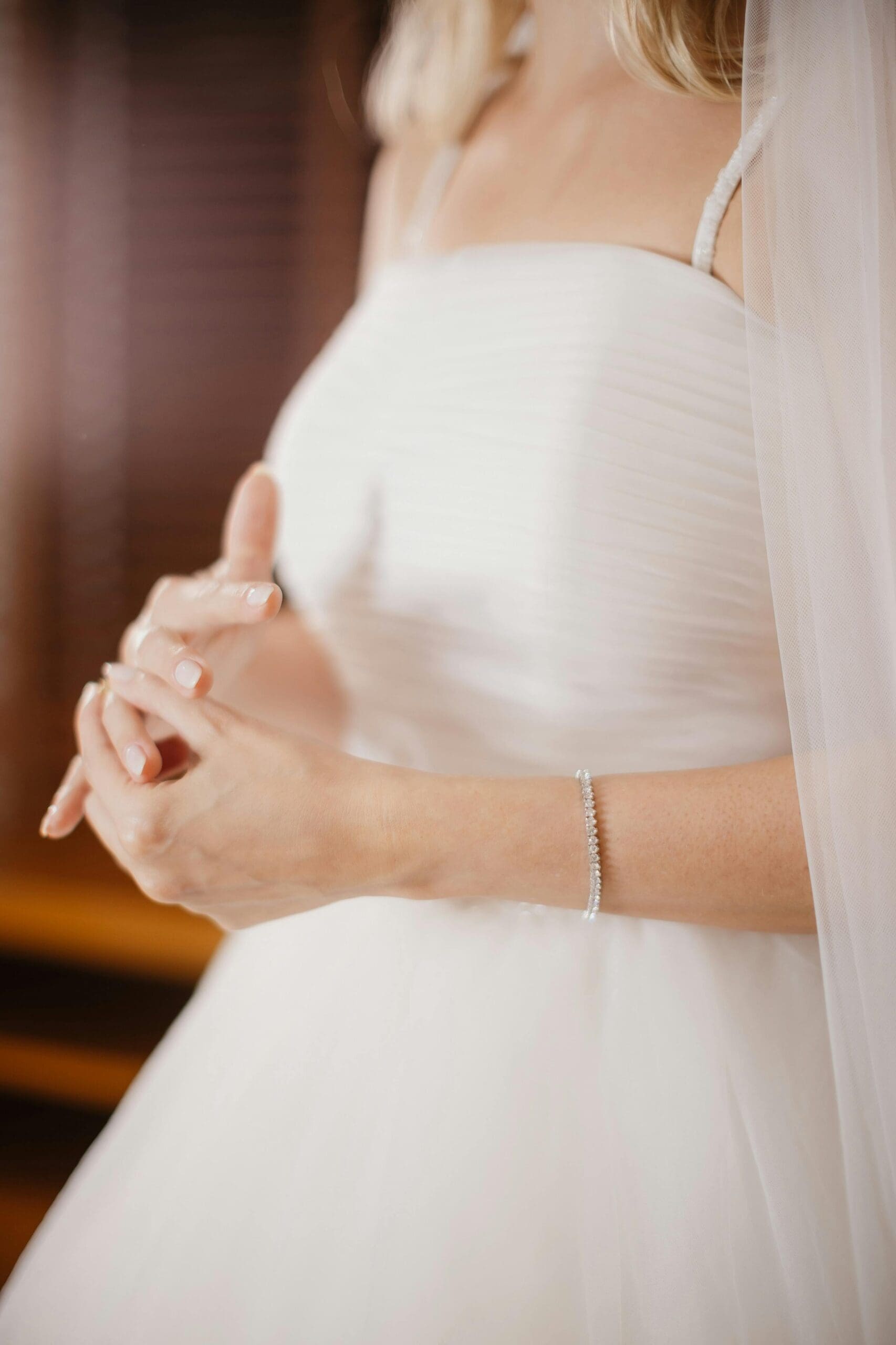A bride in a white gown and veil with hands clasped, wearing a delicate diamond bracelet, photographed in soft warm light.