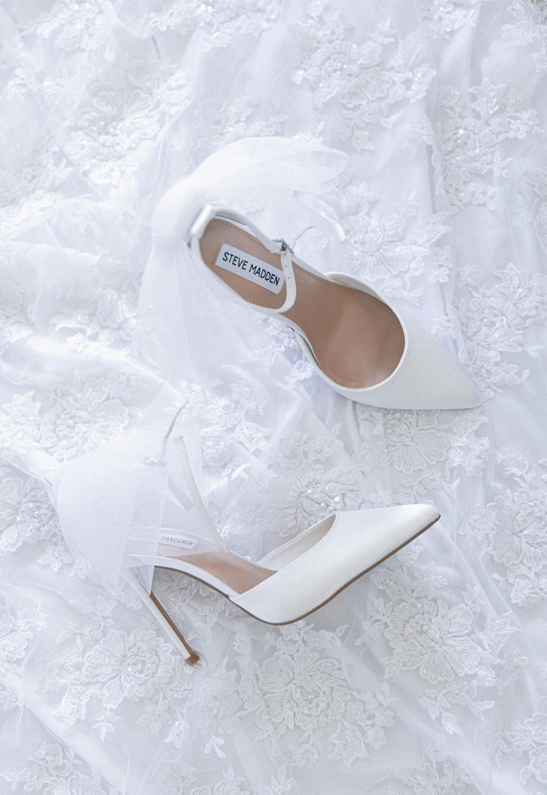 White satin bridal heels with tulle ankle straps laid on top of a lace wedding gown, styled as a flat lay detail shot.