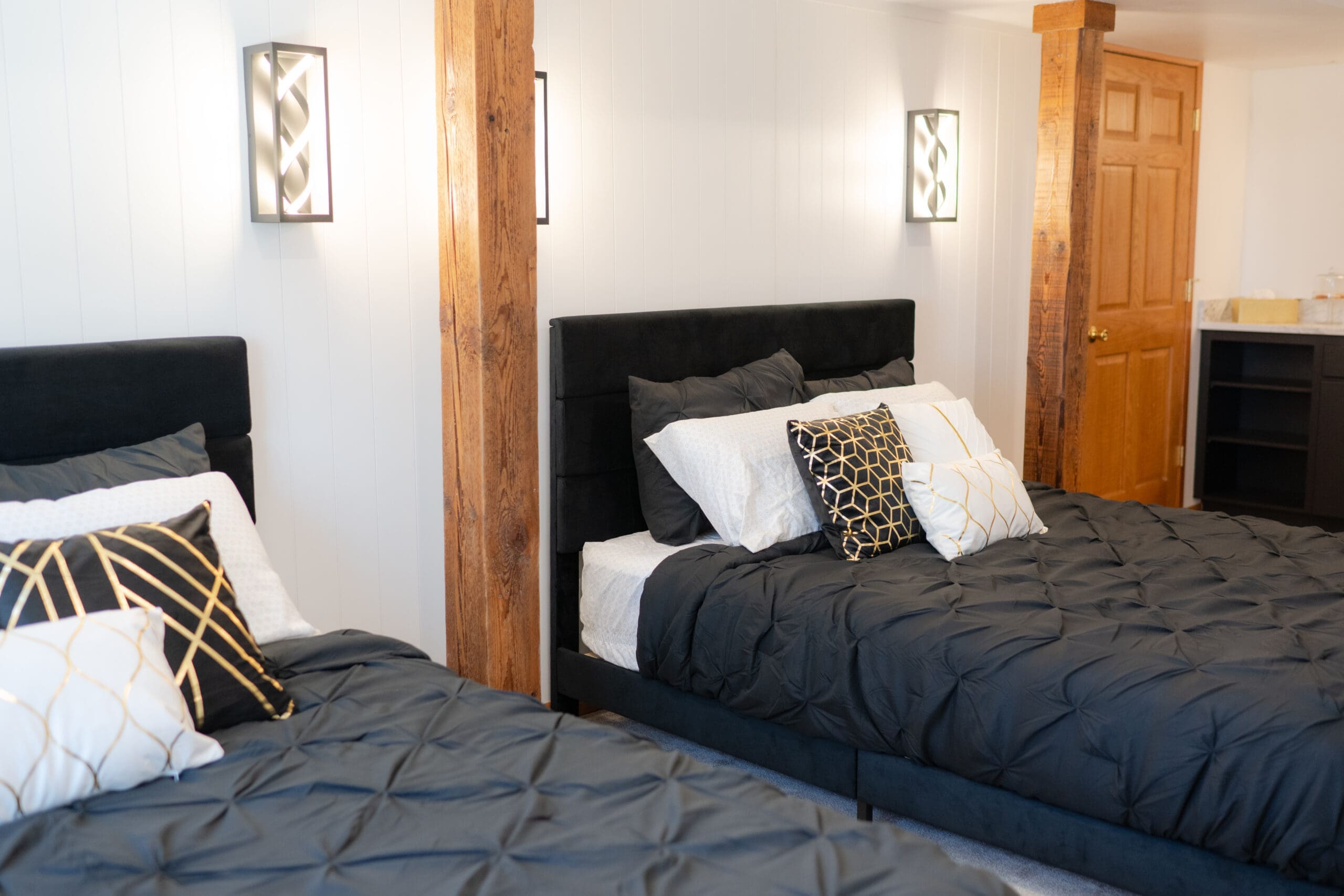 A cozy on-site wedding venue guest room at The Era in Iowa with two black upholstered beds, charcoal pintuck bedding, gold accent pillows, and rustic wood beam details.