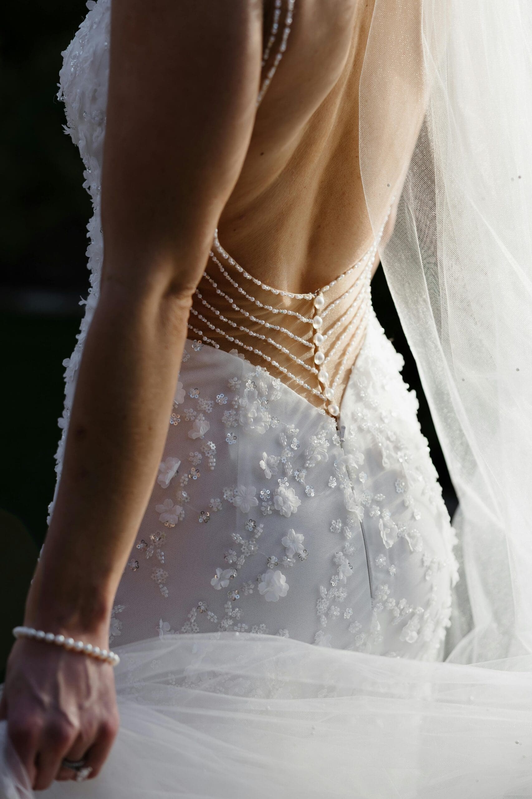 A close-up detail of a bride's embellished backless wedding gown with pearl strand accents, sequin floral appliqués, and a flowing veil at an Iowa wedding venue.