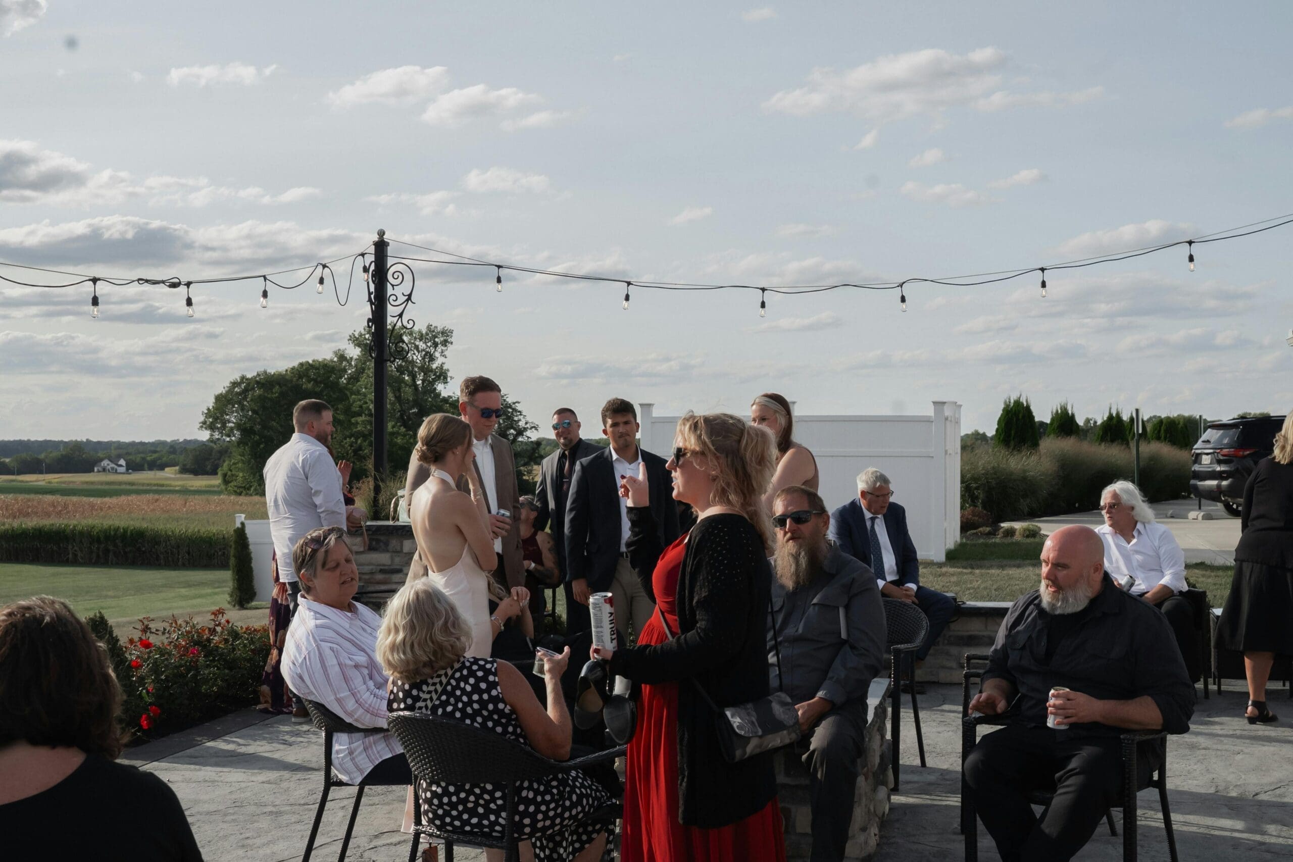 Guests mingling on an outdoor patio at a wedding venue during a rehearsal dinner, with string lights and open farmland views in the background.