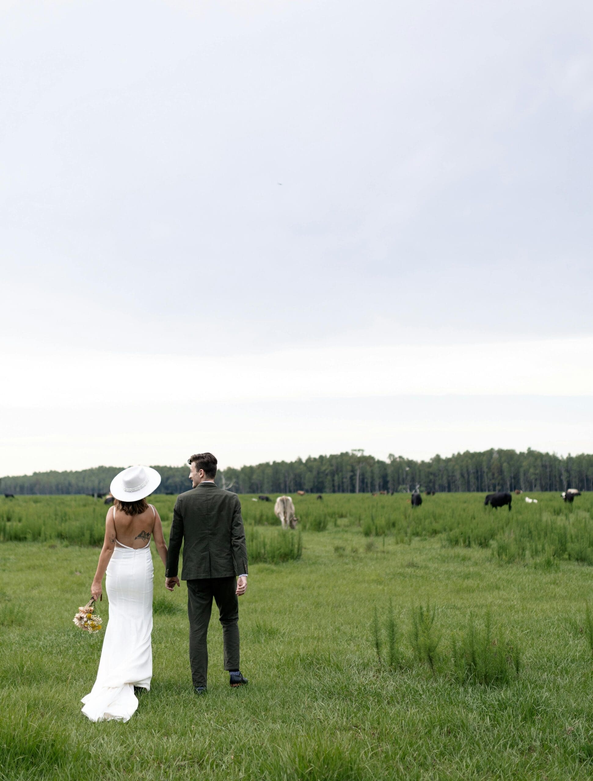 A bride in a white backless gown and wide-brim hat walking hand in hand with her groom through an open green field during outdoor wedding portraits.