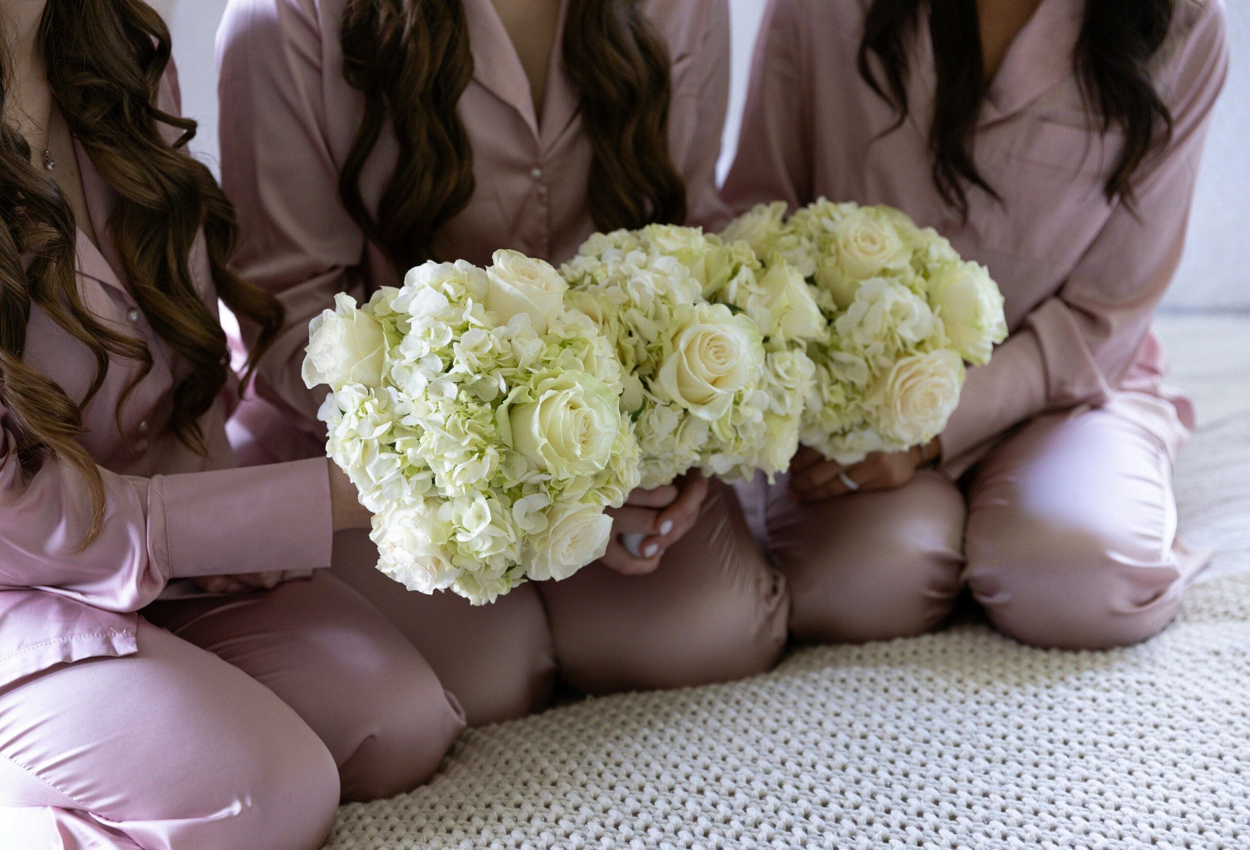 Three bridesmaids in pink satin pajamas sitting together holding lush white hydrangea and rose bouquets during wedding morning getting-ready photos.
