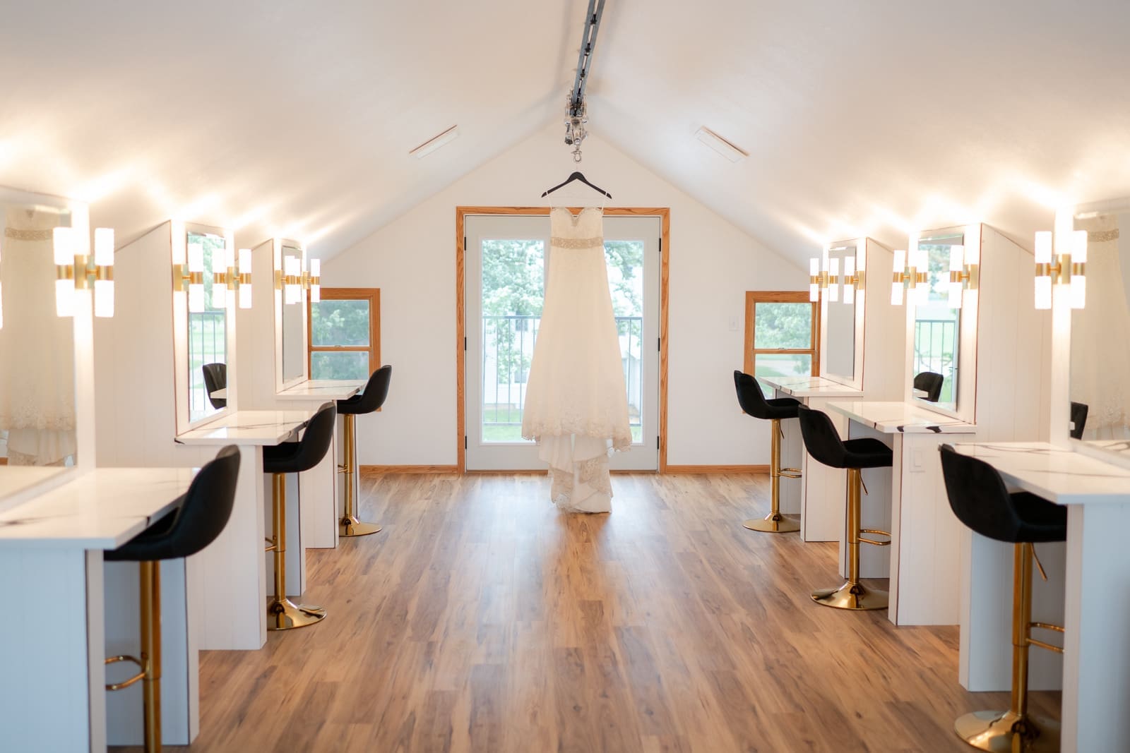 A spacious bridal getting-ready suite with six lit vanity mirrors, gold bar stools, and a wedding dress hanging in the center doorway overlooking a balcony.