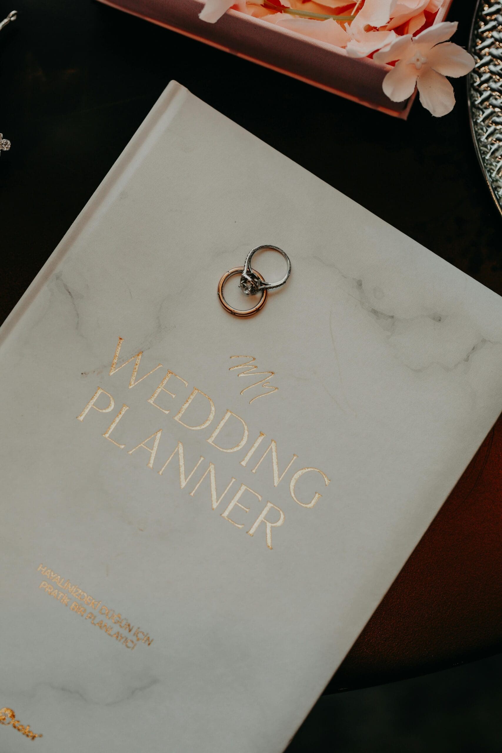 A marble and gold wedding planner book with two wedding rings resting on the cover, styled on a dark surface with pink florals in the background.