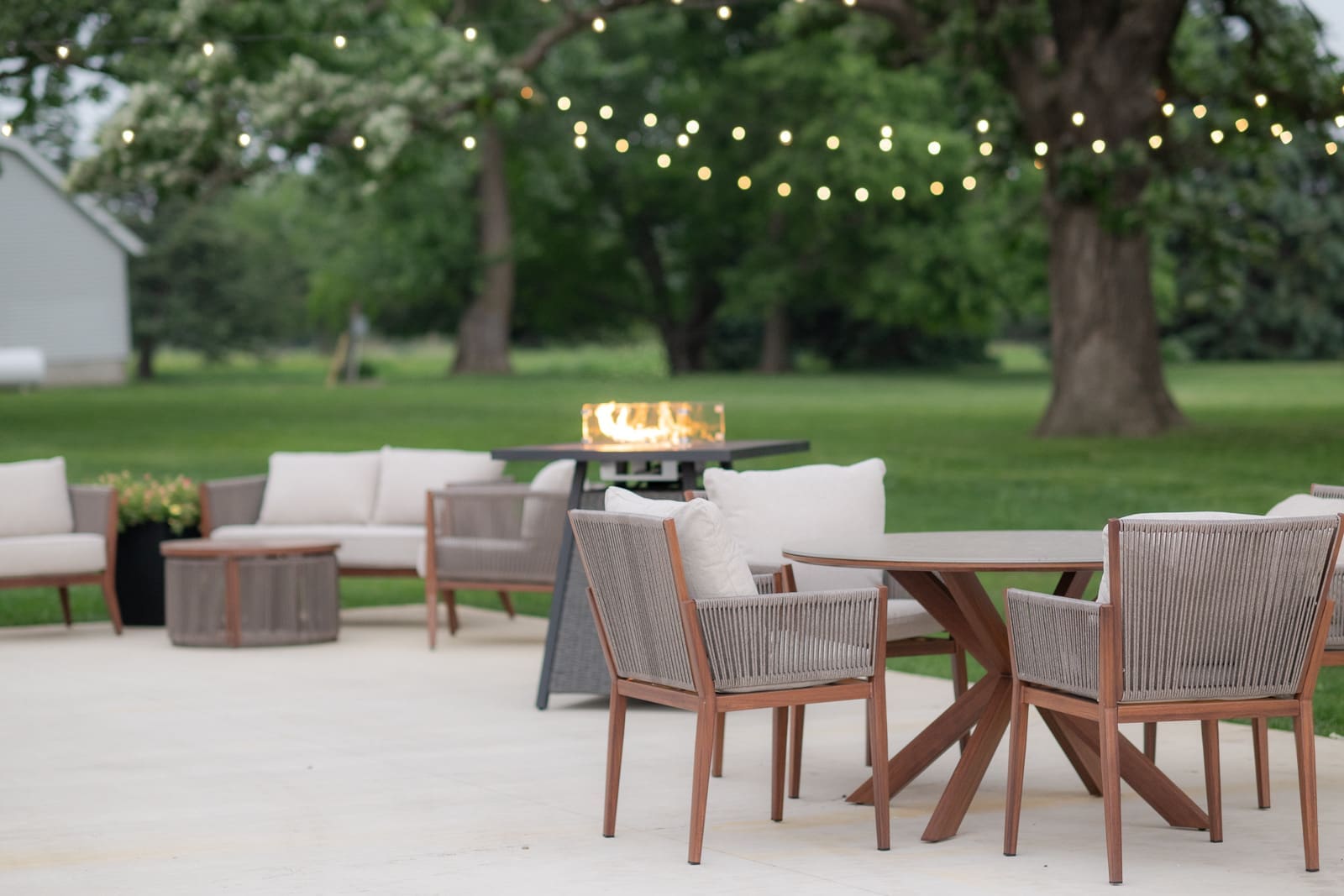 An outdoor patio at The Era wedding venue in Iowa with wicker lounge seating, a lit fire pit table, string lights, and lush green grounds at dusk.