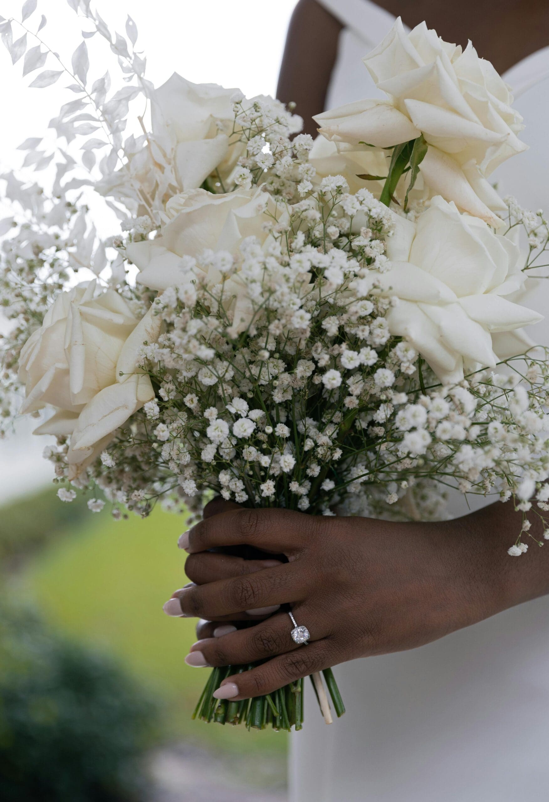 A bride holding a white rose and baby's breath bouquet with a cushion-cut diamond engagement ring visible on her hand against a soft outdoor background at her wedding weekend venue.