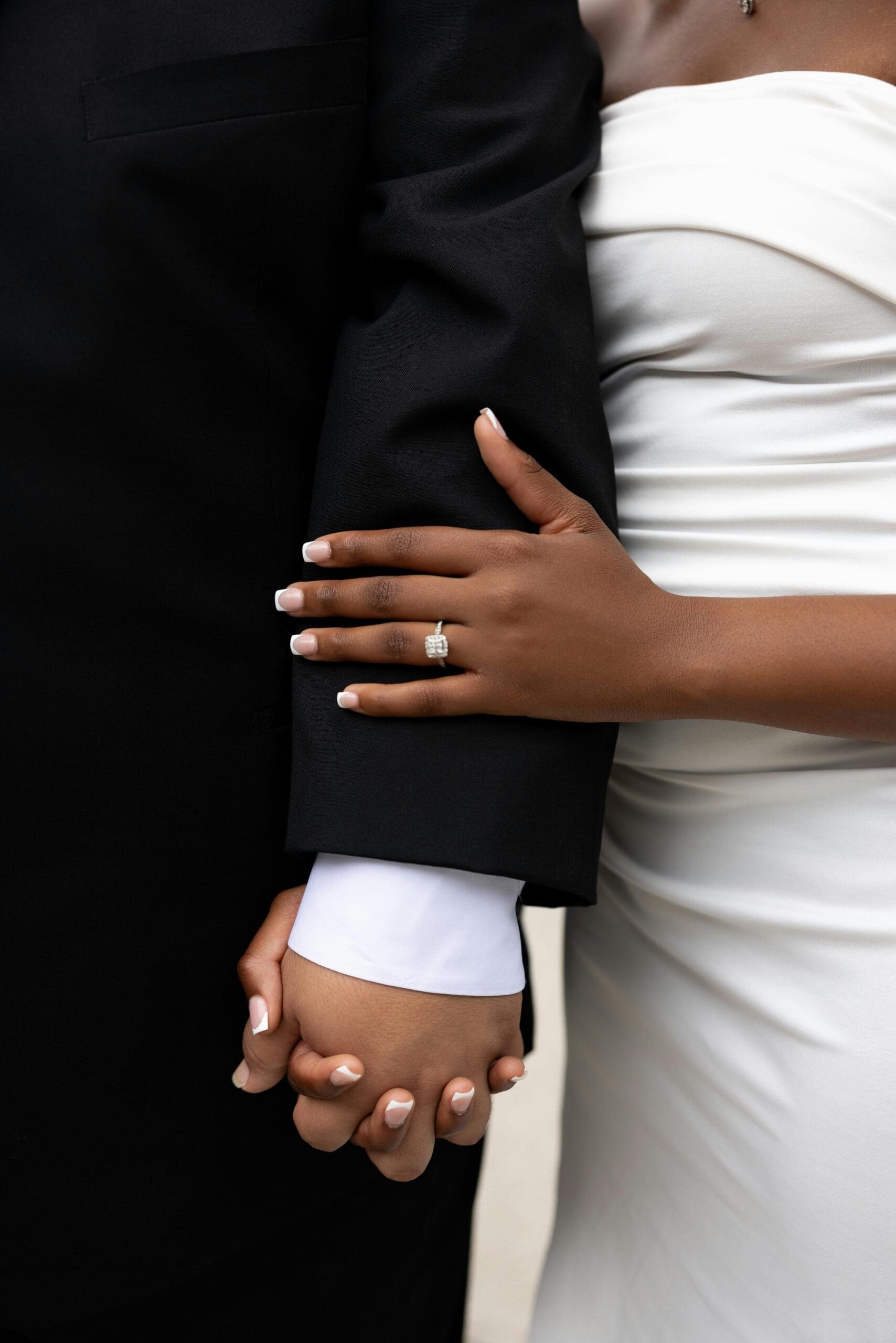 A close-up of a bride and groom holding hands, the bride's engagement ring visible as she holds her groom's arm in a white gown against his black suit.