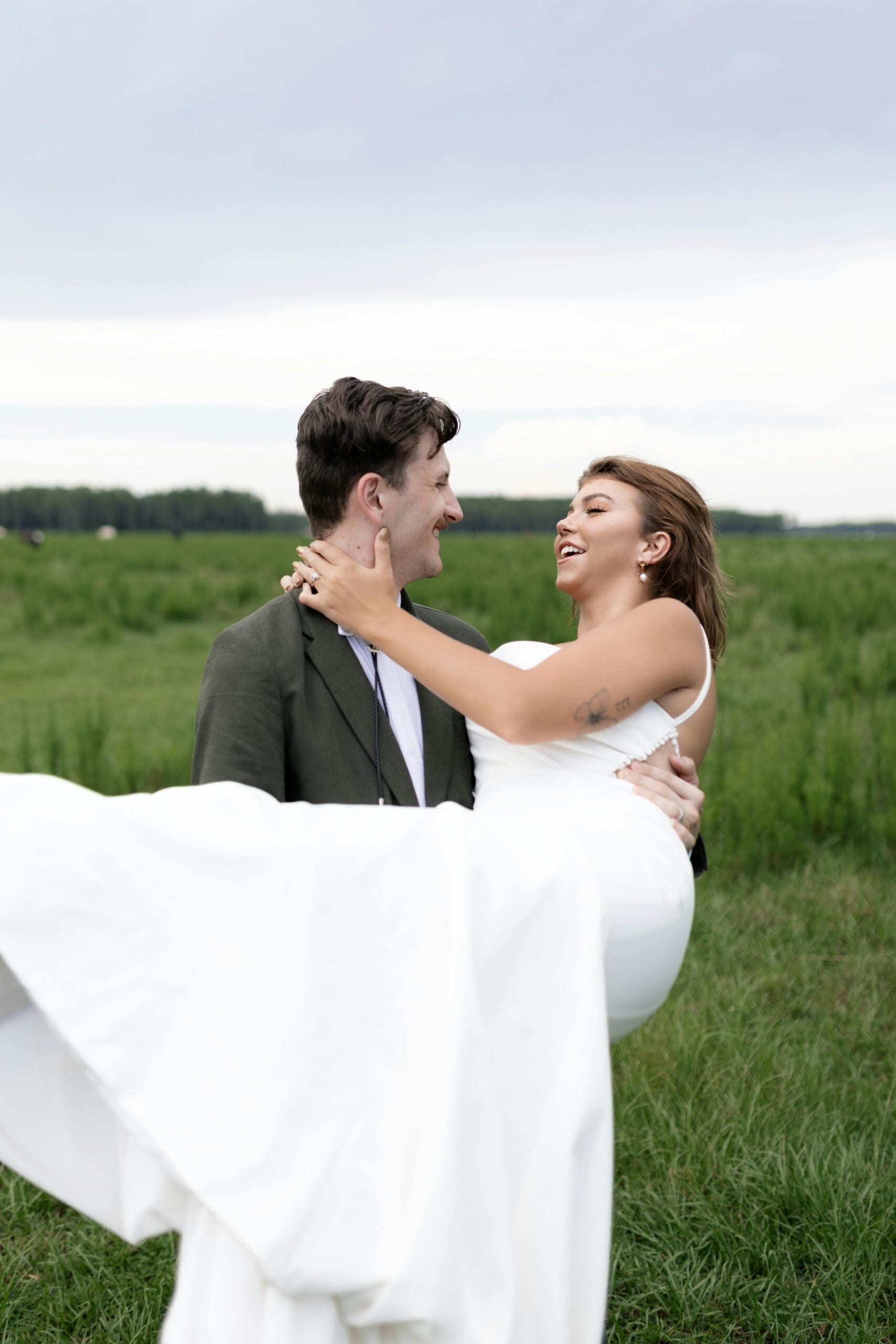 A groom in an olive suit carrying a laughing bride in a white gown through a green field during outdoor wedding portraits.
