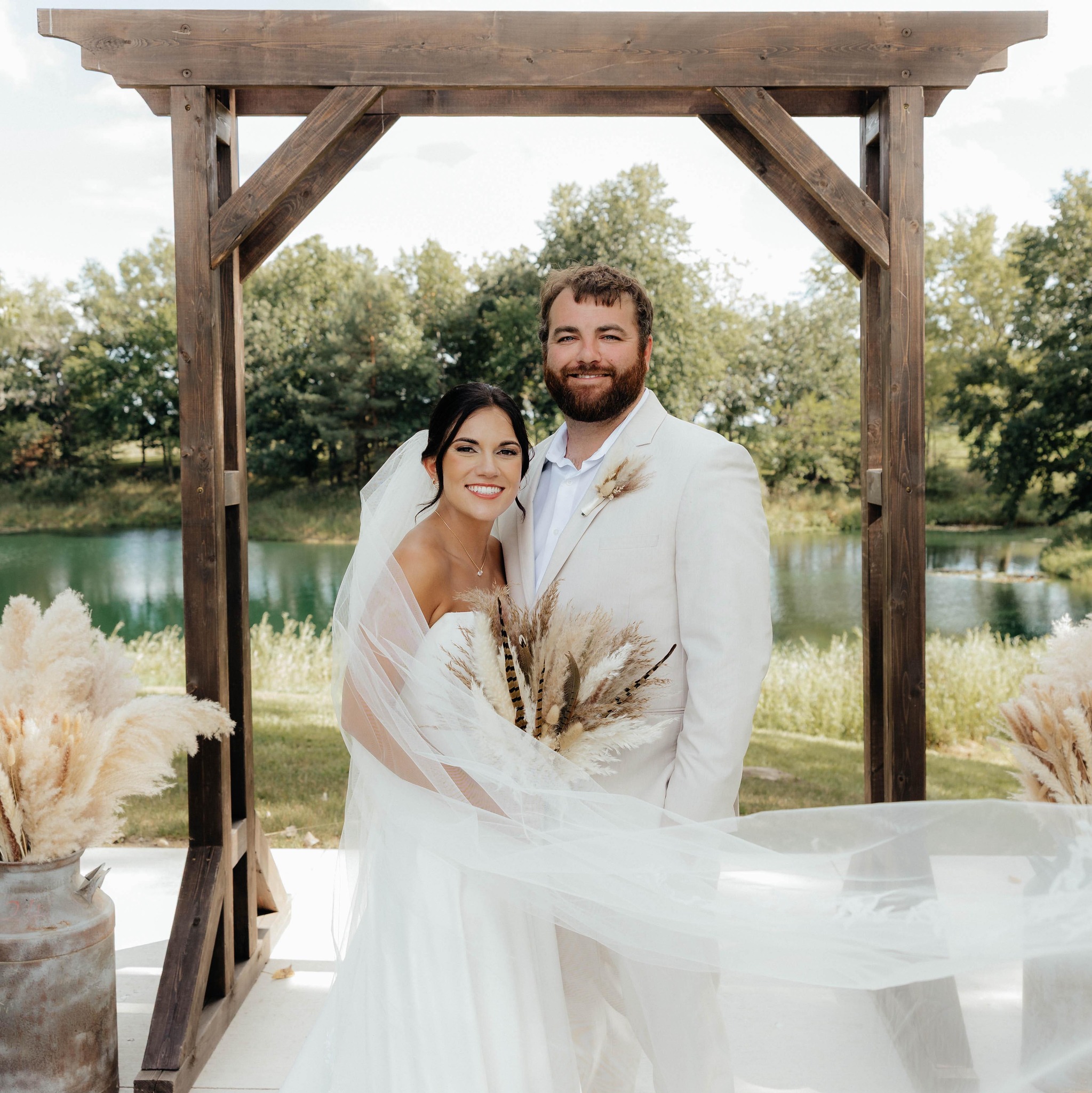 Bride and groom under a wooden ceremony arch by a pond at a country wedding venue in Iowa