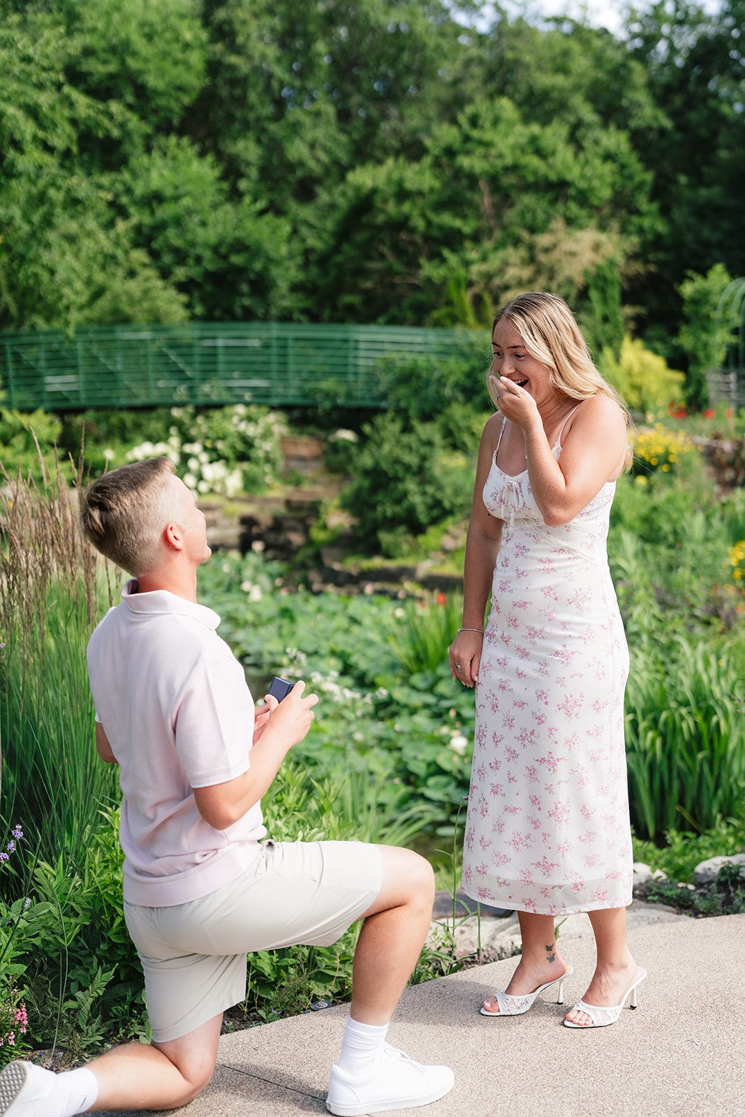 A man kneels on one knee proposing with a ring box to a smiling woman in a floral midi dress on a garden path, with a green footbridge and pond in the background.